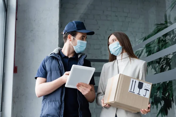 Delivery man in medical mask holding digital tablet near woman with box in hallway - foto de stock