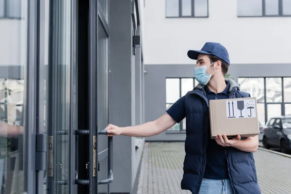 Delivery man in uniform and protective mask holding carton box and opening door of building outdoors — Stock Photo