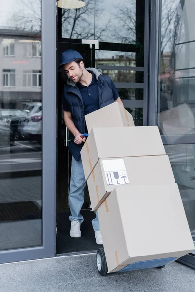 Кур'єр тримає машину з коробками біля відкритих дверей будівлі на відкритому повітрі — стокове фото