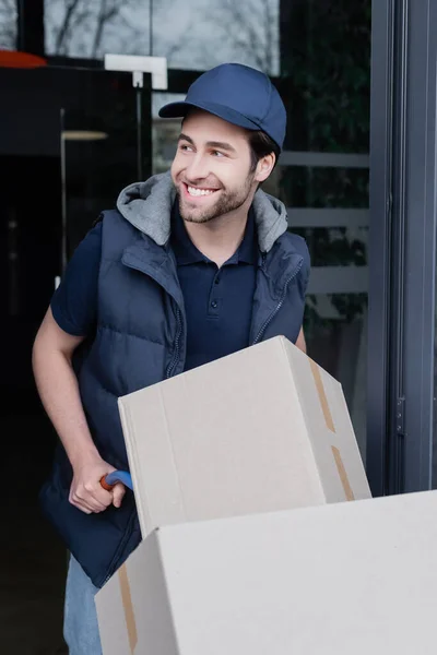 Smiling delivery man looking away near car with carton boxes and building outdoors — Stock Photo