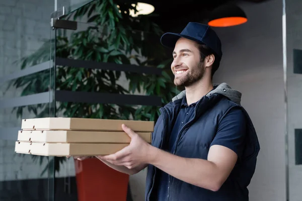 Smiling courier in uniform and cap holding pizza boxes in hallway — Stockfoto