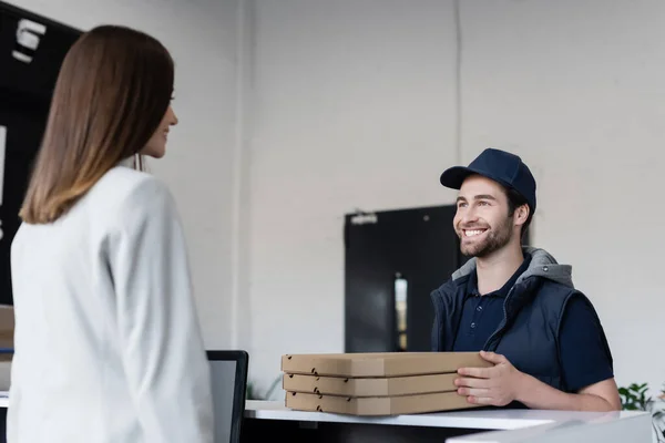 Positive courier holding pizza boxes near blurred businesswoman in office — стоковое фото