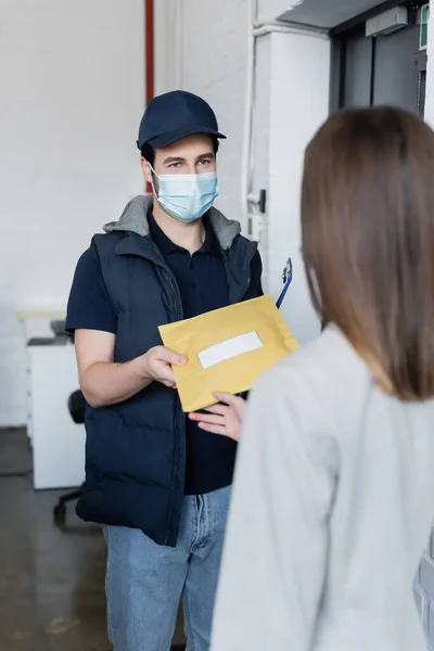 Courier in medical mask holding clipboard and giving parcel to blurred businesswoman in office — Fotografia de Stock
