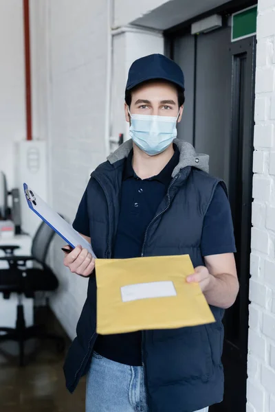 Courier in medical mask holding clipboard and parcel while looking at camera in office — стокове фото
