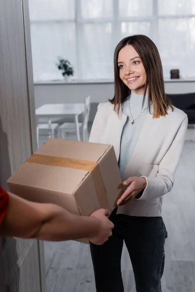 Positive woman taking carton box from blurred courier in hallway — стоковое фото