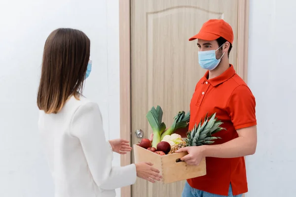 Delivery man in medical mask holding box with fresh food near woman in hallway — стоковое фото