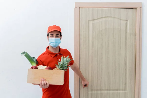 Delivery man in protective mask holding box with fresh food and looking at camera near door in hallway — Stock Photo