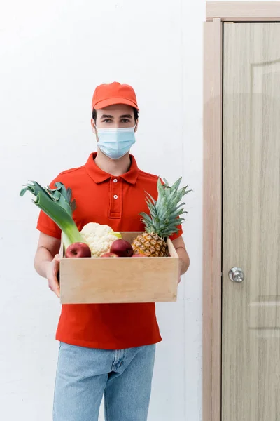Delivery man in medical mask holding wooden box with fresh food in hallway — Stockfoto