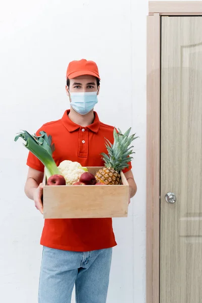 Delivery man in medical mask holding wooden box with fresh food in hallway — стоковое фото