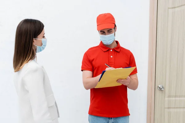 Delivery man in medical mask holding parcel and writing on clipboard near woman and door in hallway - foto de stock