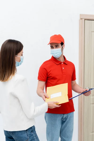 Courier in medical mask holding parcel and clipboard near customer in hallway - foto de stock