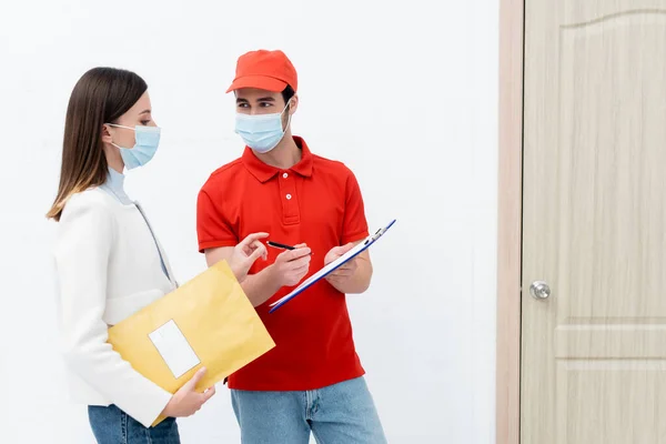 Courier holding clipboard near woman in medical mask with parcel in hallway - foto de stock