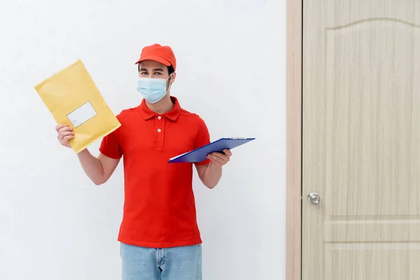 Courier in medical mask holding parcel and clipboard near door in hallway — Stock Photo