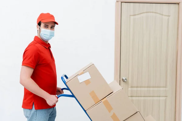 Courier in medical mask looking at camera near cart with boxes and door in hallway — стокове фото