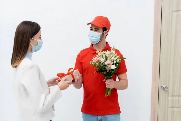 Delivery man in medical mask giving present and flowers to woman in hallway - foto de stock