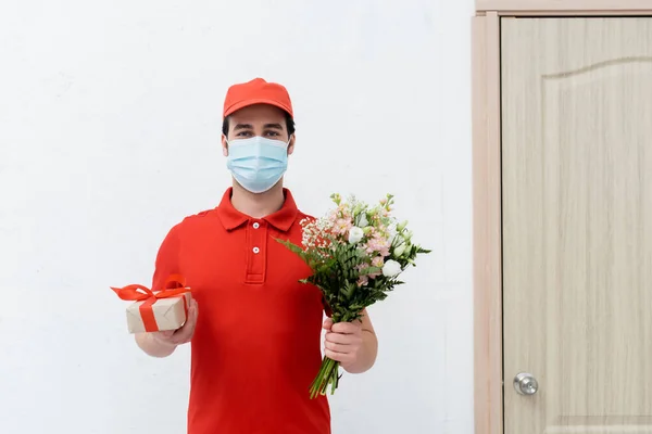 Delivery man in medical mask holding bouquet and present in hallway — Stock Photo