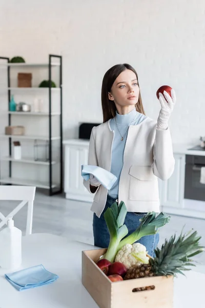 Woman in latex gloves holding rag and apple near fresh food in kitchen — стоковое фото