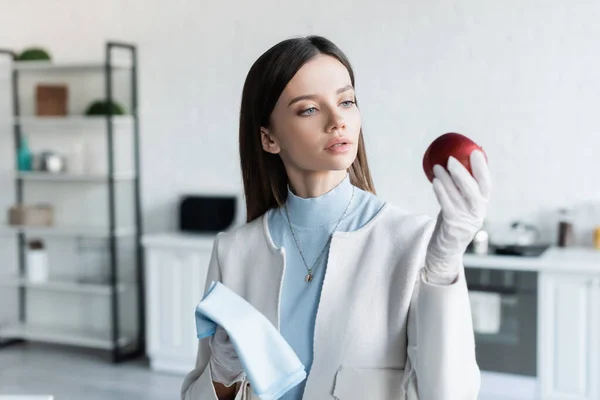 Young woman in latex gloves holding apple and rag in kitchen — Stockfoto
