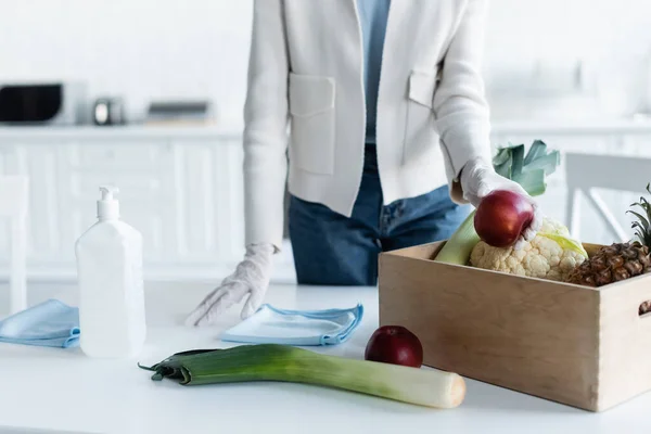 Cropped view of woman in latex gloves taking apple from box near rags and antiseptic at home — Photo de stock