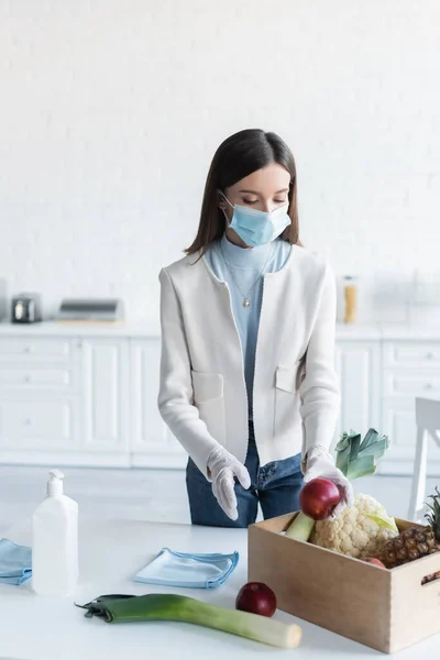 Woman in medical mask and latex gloves taking food from box near antiseptic in kitchen — Stock Photo