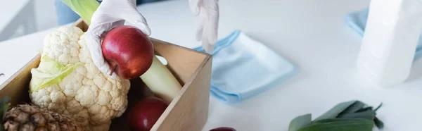 Cropped view of woman in latex gloves holding apple near food, rags and antiseptic at home, banner — стоковое фото