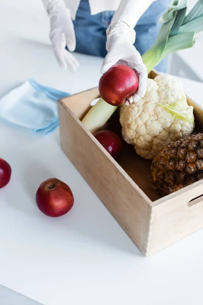 Cropped view of woman in latex gloves holding apple near wooden box with fresh food and rag at home — стоковое фото