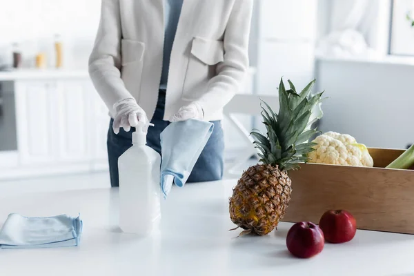 Cropped view of woman in latex gloves pouring antiseptic on rag near box with fresh food in kitchen — Stockfoto