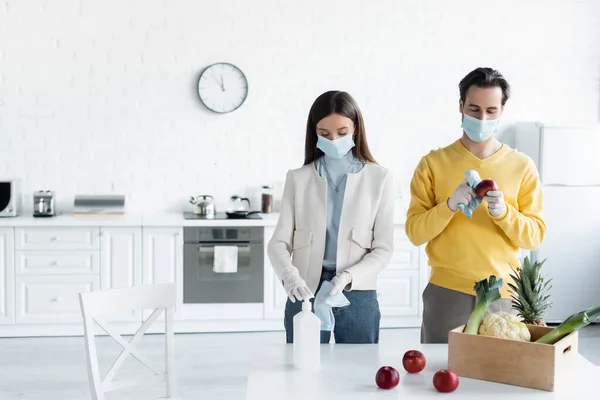 Couple in medical masks and latex gloves cleaning fresh food with antiseptic in kitchen — Photo de stock