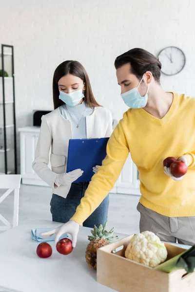 Man in medical mask holding apples near girlfriend in latex gloves holding clipboard in kitchen — стоковое фото