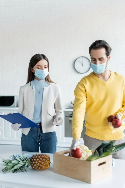 Man in latex gloves and medical mask holding apples near girlfriend with clipboard in kitchen - foto de stock