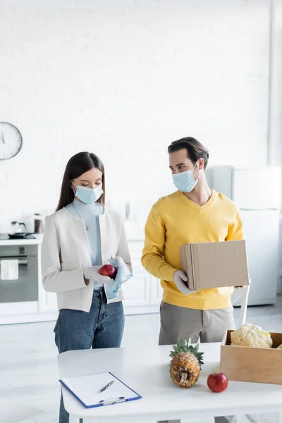 Man in medical mask holding carton box near girlfriend cleaning apple and clipboard in kitchen — Stockfoto