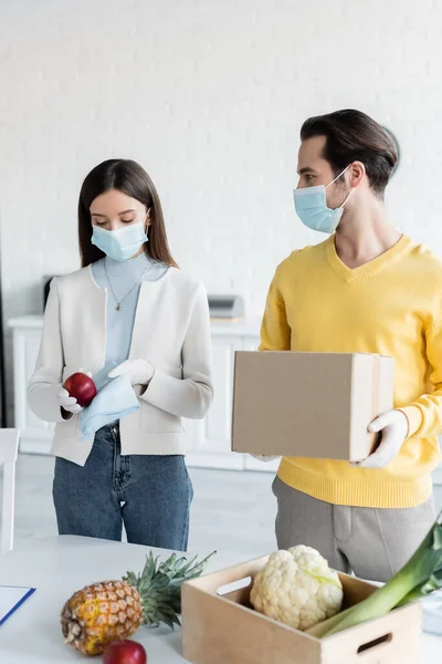 Man in medical mask holding carton box near girlfriend cleaning apple with rag at home — Stockfoto