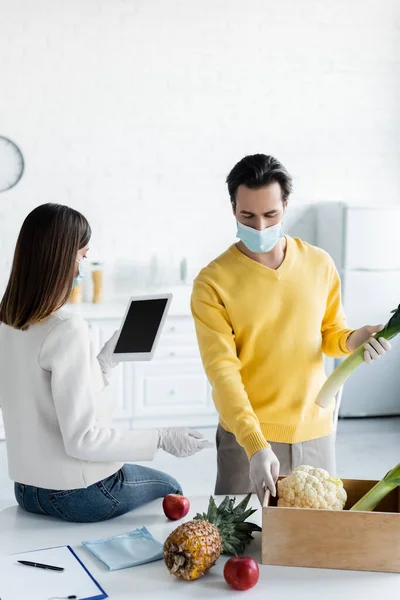 Man in medical mask and latex gloves taking food from box near girlfriend with digital tablet in kitchen — Stockfoto