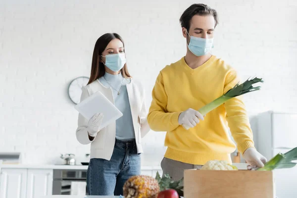 Woman in medical mask holding digital tablet near boyfriend in latex gloves taking vegetables from wooden box in kitchen — стоковое фото