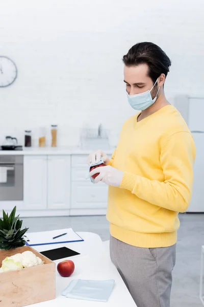 Man in medical mask and latex gloves cleaning apple with rag near fresh food and digital tablet in kitchen — стоковое фото