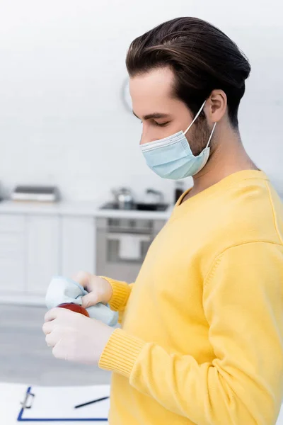 Man in medical mask and latex gloves cleaning apple with rag in kitchen — Stockfoto