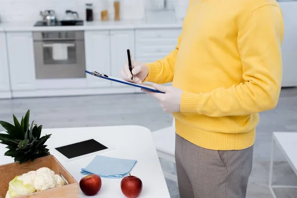 Cropped view of man in latex gloves writing on clipboard near fresh food and digital tablet in kitchen — Foto stock