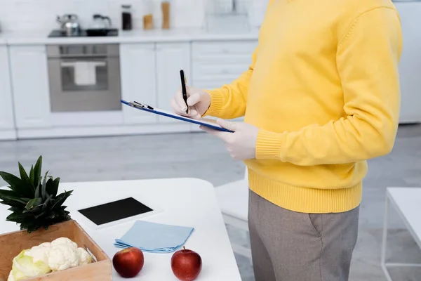 Cropped view of man in latex gloves writing on clipboard near fresh food and digital tablet in kitchen — Fotografia de Stock