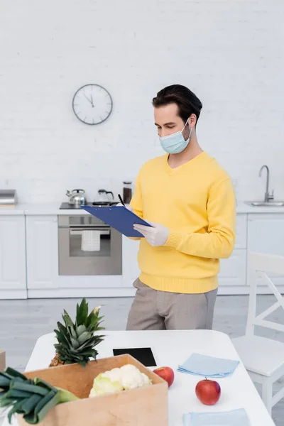 Man in medical mask and latex gloves writing on clipboard near organic food and clipboard in kitchen — Stockfoto
