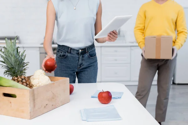 Cropped view of woman using digital tablet near fresh food and boyfriend with cardboard box at home — Photo de stock