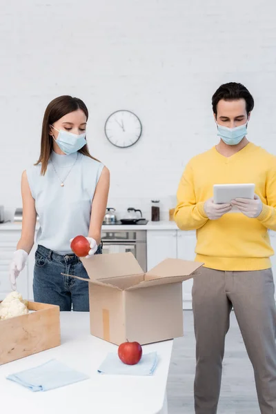 Woman in protective mask and latex gloves holding apple near boxes and boyfriend using digital tablet in kitchen — Photo de stock
