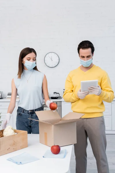 Man in latex gloves and medical mask using digital tablet near girlfriend, fresh food and boxes in kitchen — Stockfoto