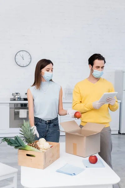 Man in medical mask using digital tablet near girlfriend holding apple and food in boxes in kitchen — Photo de stock
