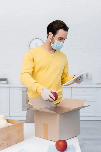 Man in medical mask and latex gloves using digital tablet and holding apple near carton box at home — Photo de stock