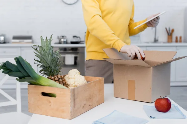 Cropped view of man in latex gloves using digital tablet and taking apple from carton box in kitchen — Stock Photo