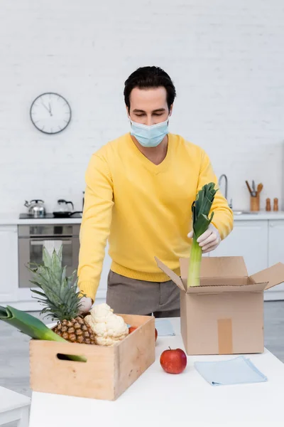 Young man in medical mask holding leek near fresh food and rags at home - foto de stock