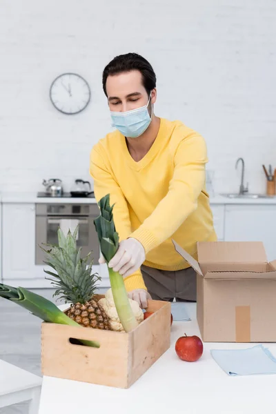 Man in protective mask taking leek from box with food near rags in kitchen — стоковое фото