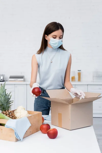 Woman in latex gloves and medical mask taking apple from carton box near rags in kitchen — стоковое фото