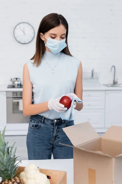 Woman in medical mask cleaning apple near boxes in kitchen — Stock Photo