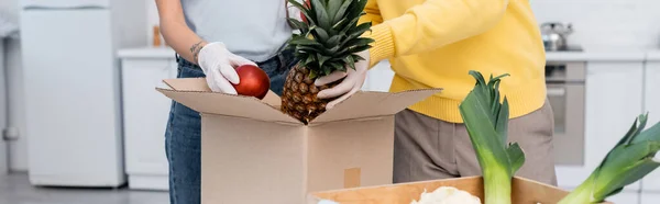 Cropped view of couple in latex gloves taking fruits from box in kitchen, banner — стоковое фото